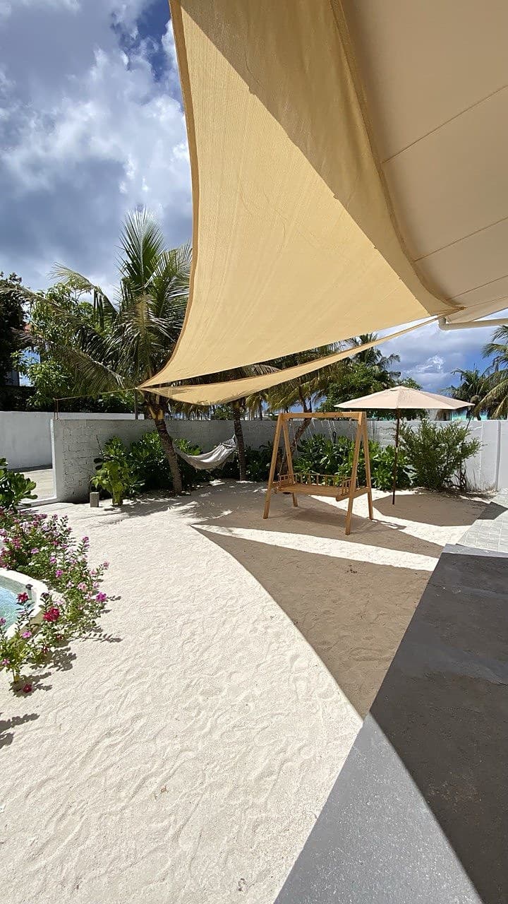 A shaded garden lounge under a beige shade sail with a wooden swing seat, hammock and flowering plants beside the plunge pool